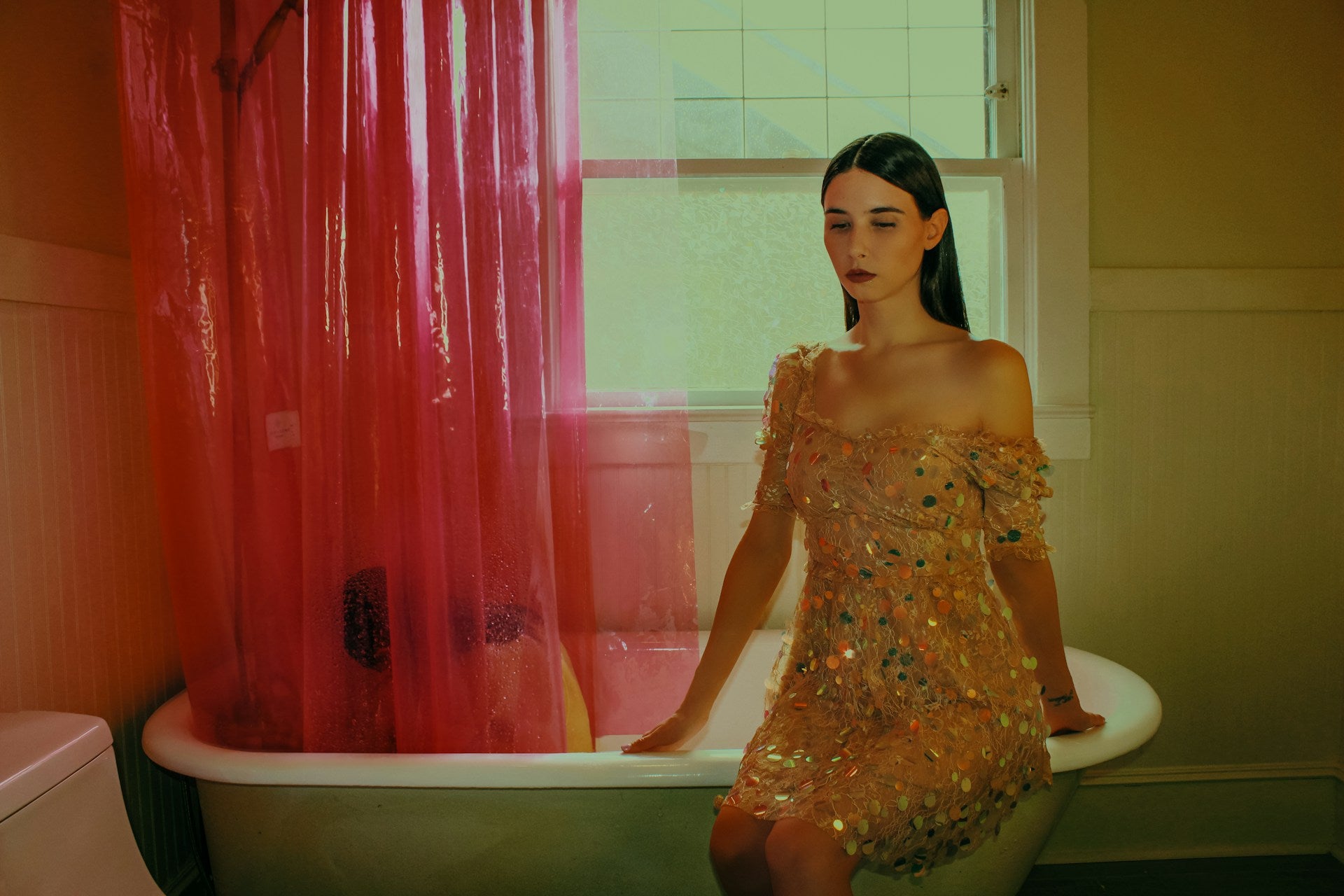 Woman in a light-colored dress decorated with colorful stones sits on the edge of a bathtub. The red shower curtain is still slightly wet.