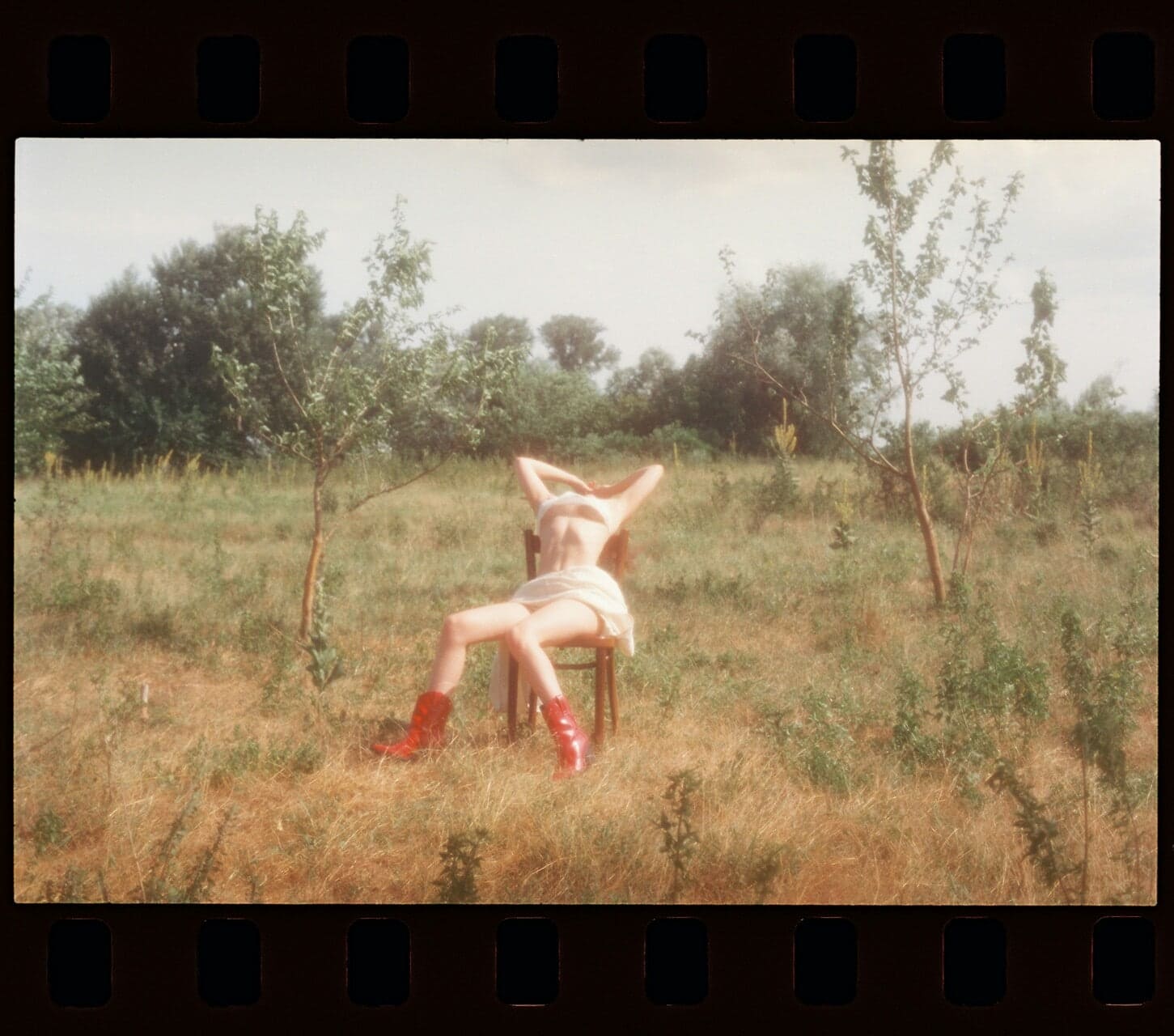 Woman in red boots sits on a wooden chair outdoors and leans back over the chair. Filmed on film reel. Scene appears artistic.