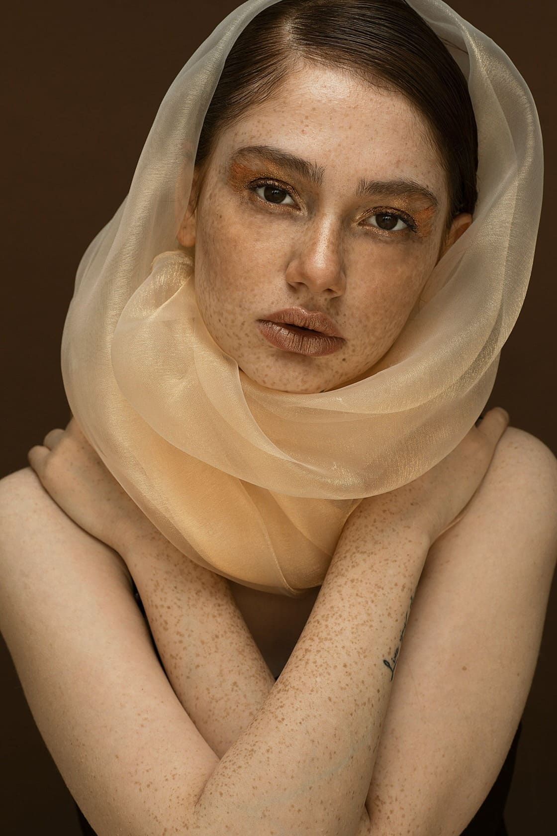 Portrait of a fair-skinned, red-haired woman. The woman has a very light veil around her head, her freckled arms crossed over her shoulders, and she is looking directly into the camera.