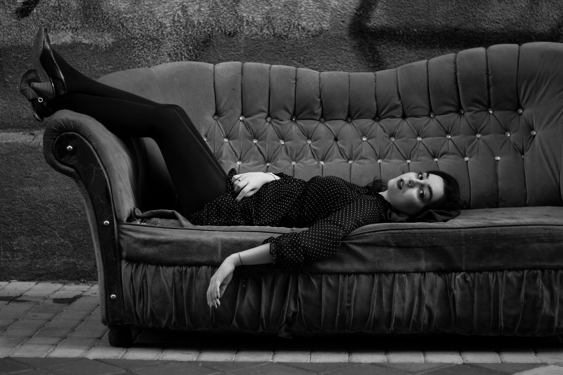 Hellhäutige Frau in gepunkteter Bluse und schwarzer Leggings liegt mit dem Rücken auf einer Couch und schaut direkt in die Kamera. Couch steht auf der Straße vor einer Wand mit Grafitis. Schwarz-weiß Fotografie. Szene wirkt ästhetisch.