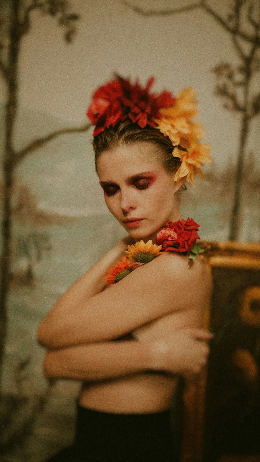 Topless woman wraps her arms around her upper body. Flowers in her hair and on her shoulder. Eyes painted red. Half-body portrait. Scene appears artistic.