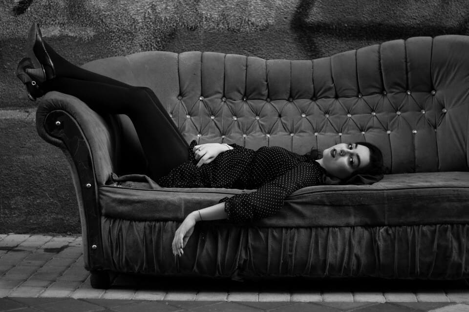 Una mujer de piel clara con una blusa de lunares y leggings negros está tumbada boca arriba en un sofá y mira directamente a la cámara. El sofá está en la calle, delante de una pared con grafitis. Fotografía en blanco y negro. La escena tiene un efecto estético.