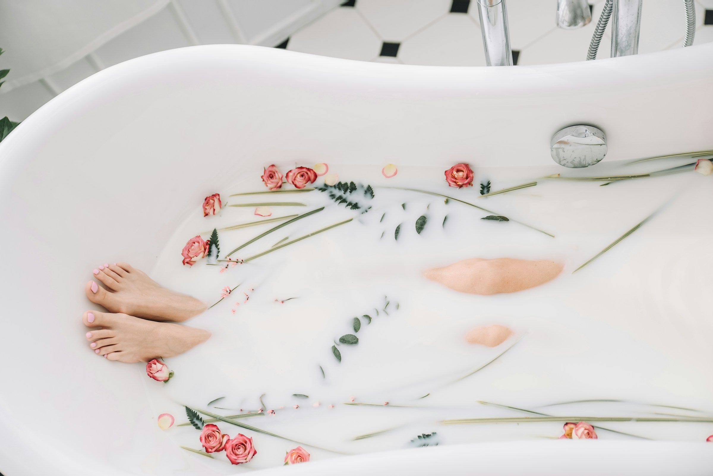 Woman relaxing in a white bathtub filled with milky water, surrounded by red rose petals and green leaves