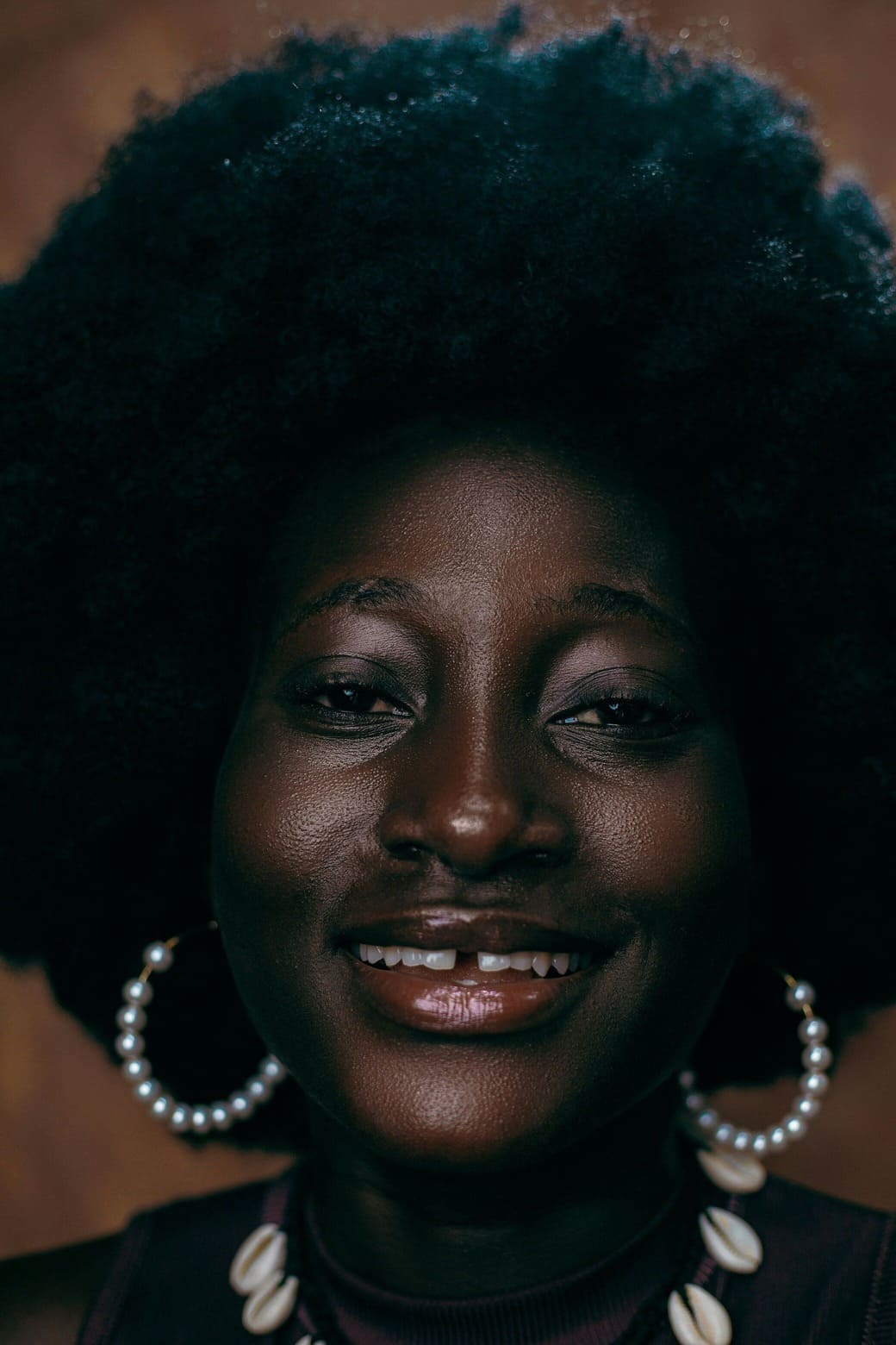 Una mujer de piel oscura sonríe a la cámara en un retrato. Tiene un poco de purpurina en la cara y lleva pendientes de perlas blancas.