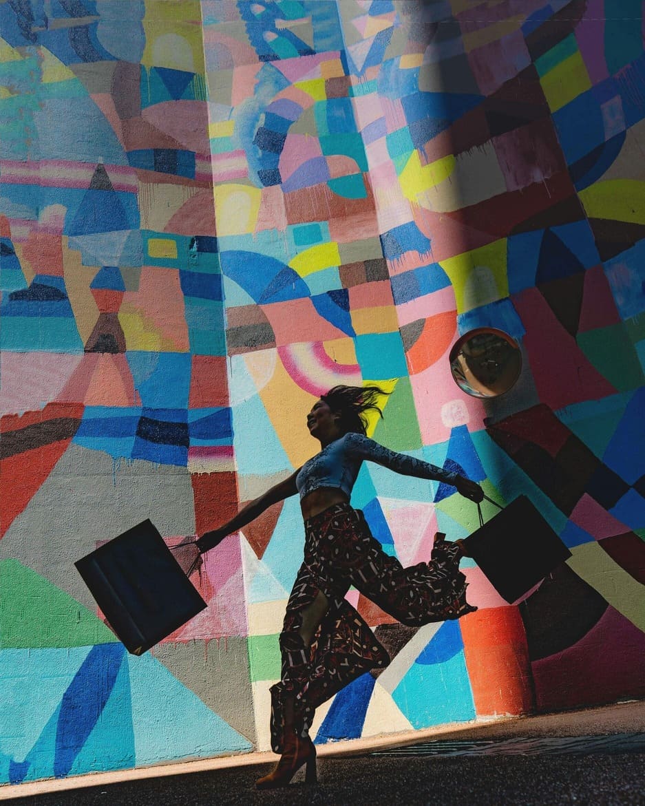 Happy woman with two shopping bags and a colorful outfit dances in front of a large, colorful wall.