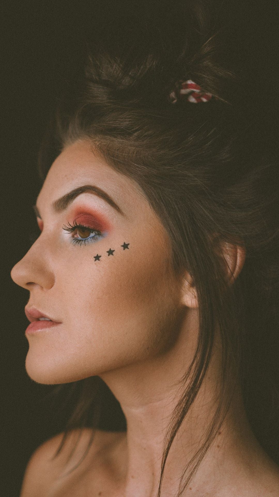 Fair-skinned, dark-haired woman with red makeup and three stars painted on her face looks to the side. Portrait photography.