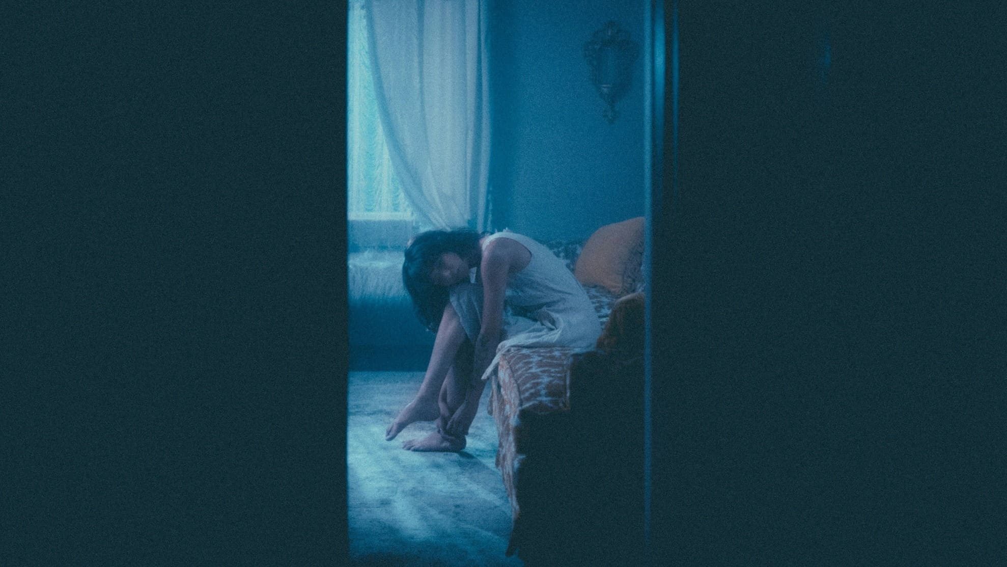 Asian woman sits on a couch and leans her upper body dreamily on her leg. She appears dreamy. View through a crack in the door.