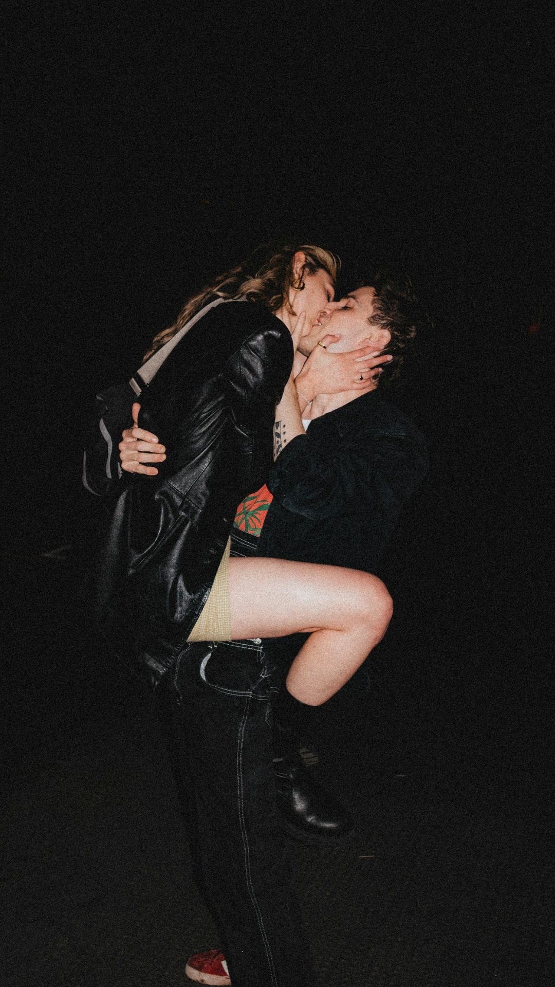 Pareja joven vestida de negro besándose. La mujer se ha subido encima del hombre, que está de pie y la sostiene en el aire. La escena parece ser de una salida nocturna.