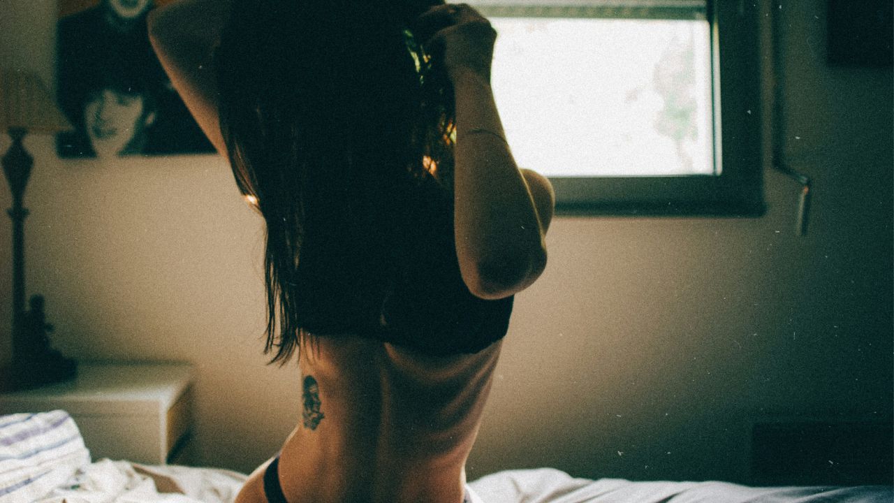 A thin, athletic, dark-haired woman sits on the bed in her underwear and runs her fingers through her hair. The scene has a documentary feel to it.