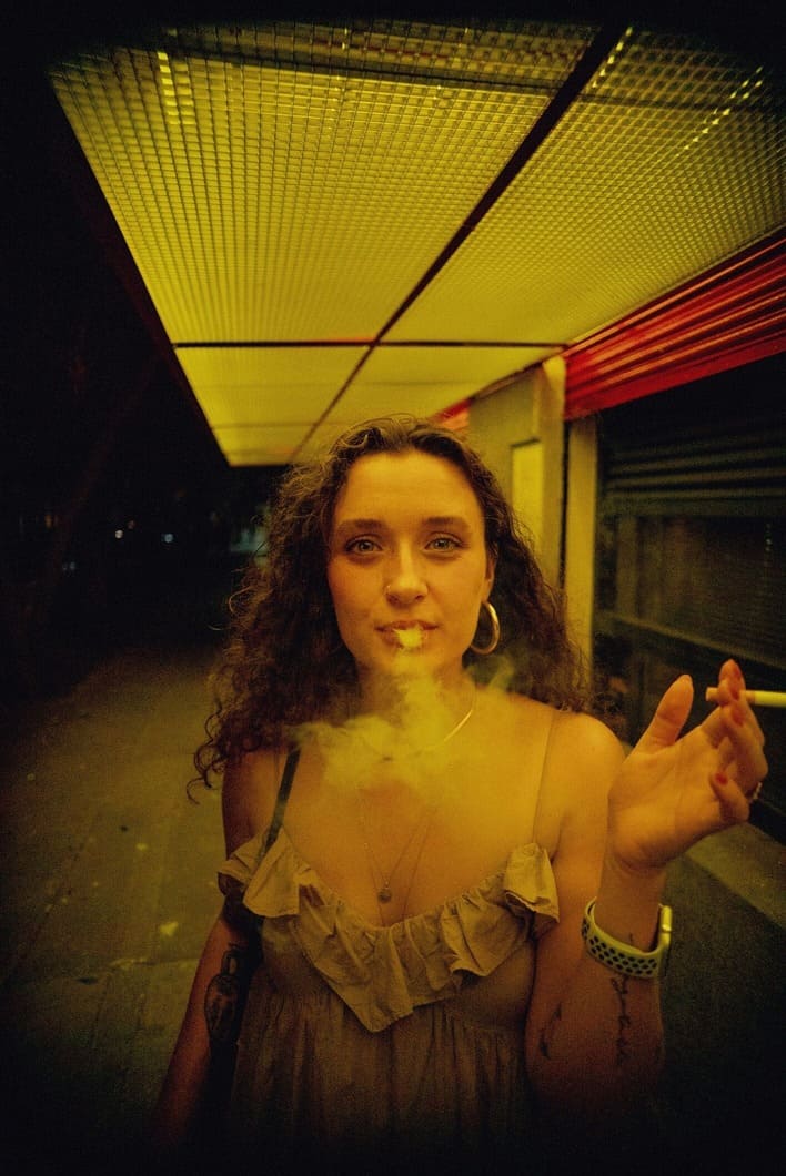 A curly-haired, fair-skinned woman smokes a cigarette and looks into the camera. She is standing in front of an empty store in a dark environment, wearing a dress and carrying a handbag.