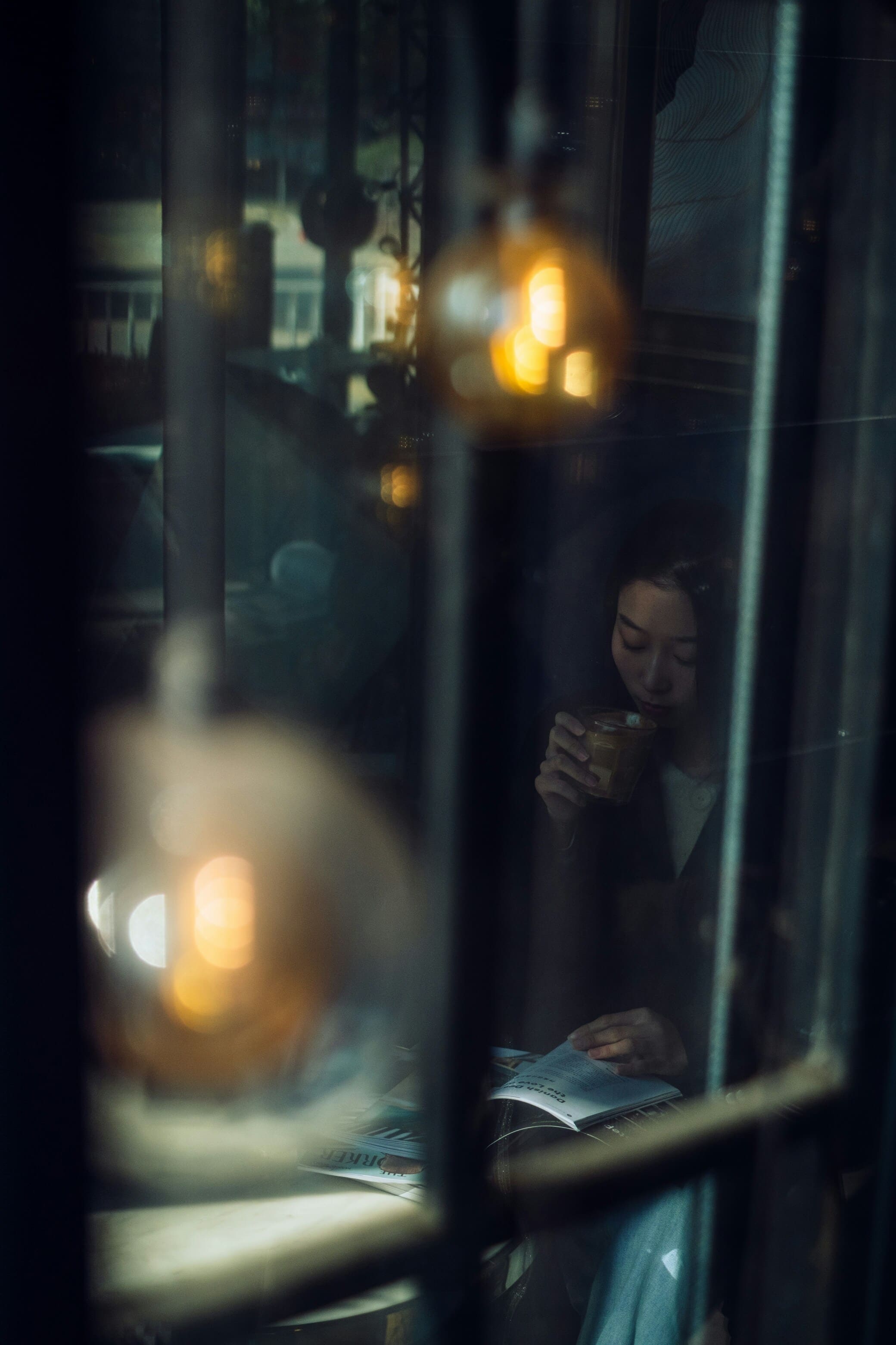 Asian-looking woman reading a book and drinking coffee. View from outside looking in through blurred glass. Incandescent bulbs illuminate the scene with yellow-orange light. Woman is rather indistinct.