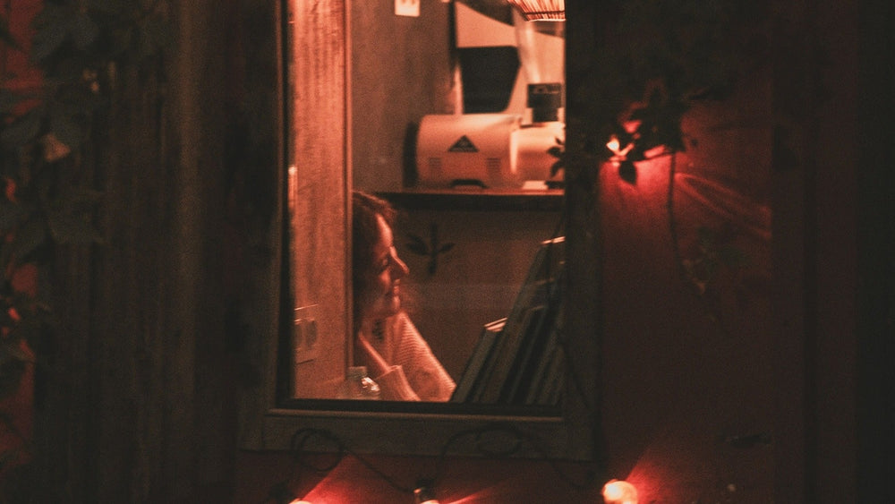 A woman is sitting in her apartment, smiling. View from outside through an open window. Red light illuminates the scene.
