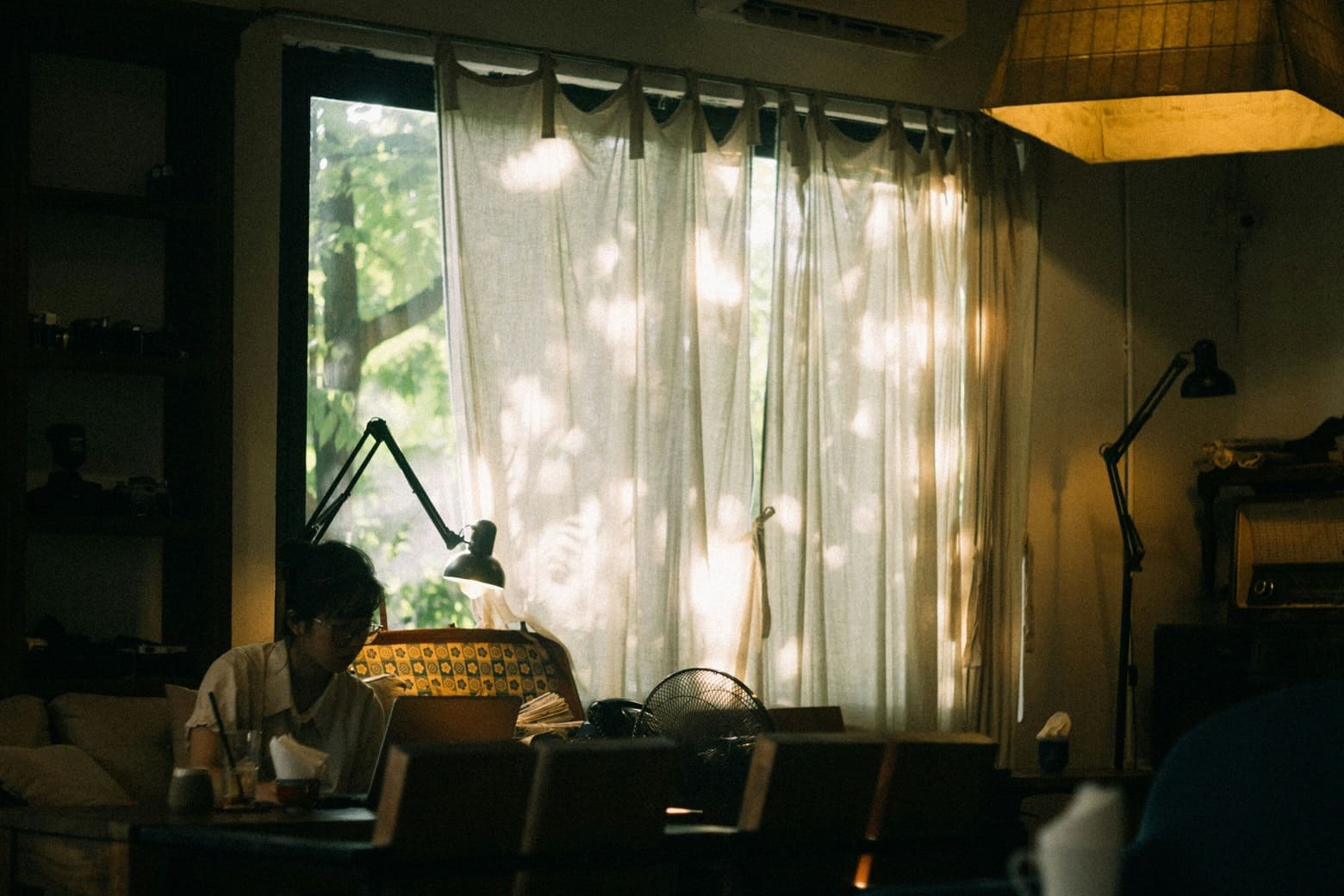 Asiatische Frau sitzt in ihrem Wohnzimmer. Gelbes Licht von oben und Tageslicht scheint durch ein großes Fenster. Die Frau sitzt am Tisch vor dem Laptop. Szene wirkt dokumentarisch.